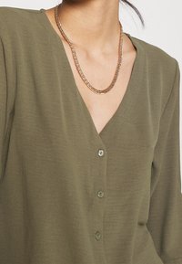 Blouse vert olive avec un col en V, manches courtes et boutons sur le devant. Portée avec un collier en chaîne dorée. Texture de tissu douce.