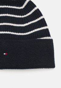 Strikket lue med marineblå og hvite horisontale striper. Stoffet er ribbet; har en liten logo i rødt, hvitt og blått på vrangborden.