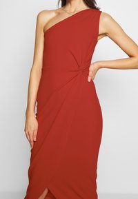 Enaxelad, figursydd klänning i mörkröd tyg med knutdetalj vid midjan och en sidofåra. Slät yta, minimal design.