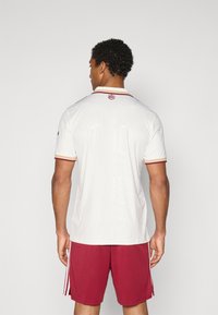 Polo branco de manga curta com padrão texturizado, realçado por uma gola e acabamentos nas mangas em borgonha, combinado com calções vermelhos com riscas laterais.