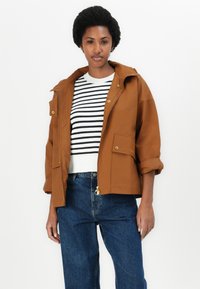 Veste à capuche marron avec des accessoires dorés, deux poches avant, portée sur un pull rayé blanc et noir et un jean bleu. Texture lisse.