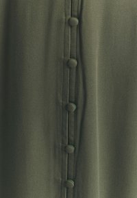 Khaki zelená látka s hladkou texturou, s centrální linií pěti knoflíků potažených látkou a jemnými šicími detaily.