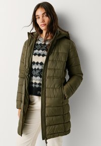 Olive zaļš polsterēts mētelis ar rāvējslēdzēju un kapuci, ar kviltētu tekstūru. Valkāts pār rakstainu džemperi un krēmkrāsas biksēm.