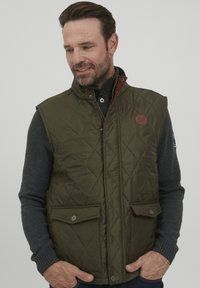 Padded zelená vesta s vysokým límcem, hn�ědými akcenty, předním zapínáním na cvoky a dvěma náprsními kapsami. Nositelná přes tmavý svetr.