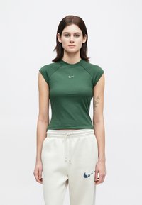 Νεαρή γυναίκα με ίσια καστανά μαλλιά, φορώντας μια πράσινη αμάνικη μπλούζα Nike και λευκές φόρμες Nike, standing against a plain background.