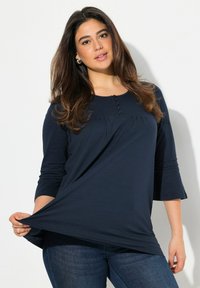 Maglia a maniche lunghe blu navy con scollo rotondo, dettagli con bottoni e vestibilità ampia, abbinata a jeans in denim scuro. Tessuto morbido con una leggera caduta.