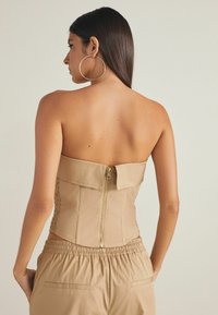 Beige strapless korsettopp med en slät yta, med en guld dragkedja bak och detalj med rynkningar på sidan. Matchas med byxor med elastisk midja.