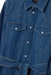 Chemise en denim de couleur bleu profond avec des boutons-pression en argent, col pointu, deux poches poitrine et une ceinture nouée à la taille.