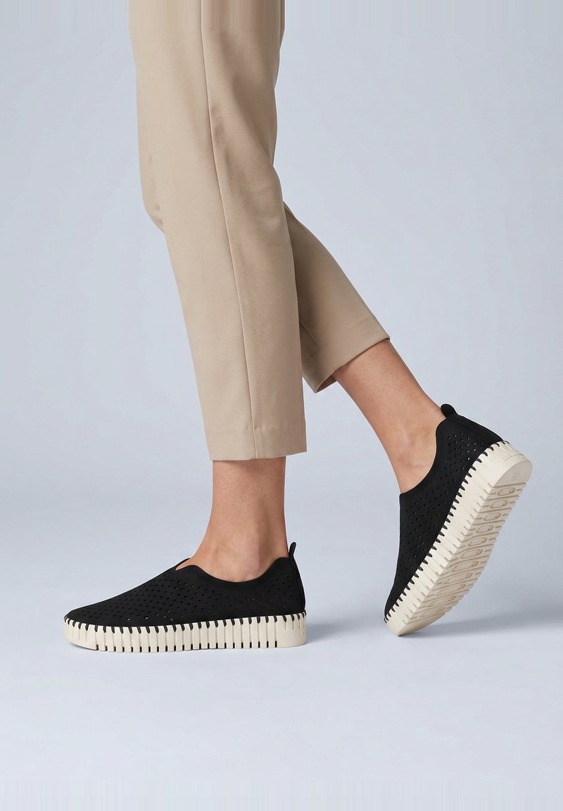 Personne portant un pantalon beige raccourci et des chaussures noires à enfiler avec des semelles blanches striées, un pied légèrement levé sur un fond uni.