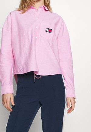 Personne portant une chemise rose clair boutonnée avec un patch logo Tommy Jeans et un pantalon bleu marine avec des bandes blanches sur les côtés.