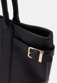 Sac à main en cuir noir avec double poignée et une lanière latérale ornée d'une boucle rectangulaire en or, présenté sur un fond clair uni.