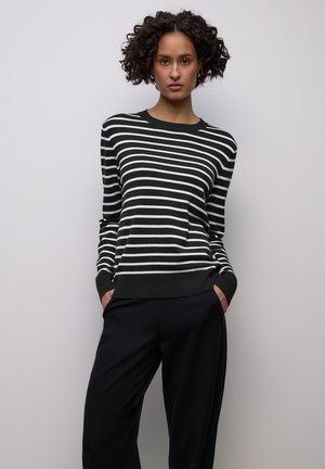 Mustavalkoraidallinen neulepusero, jossa pyöreä pääntie, resorit hihansuissa ja helmassa. Malli pukeutunut rentoihin mustiin housuihin, kädet taskuissa.