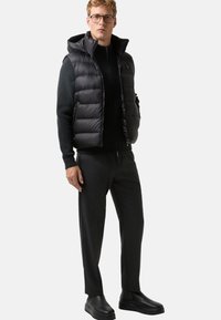 Gilet matelassé noir avec capuche sur un pull noir, associé à un pantalon sombre et des chaussures noires. Texture lisse, design épuré et style minimaliste.