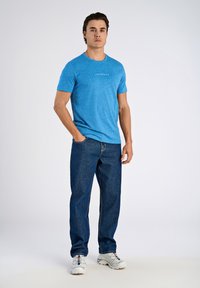 Sininen lyhythihainen t-paita, jossa "LINDBERGH"-logo, yhdistettynä sinisiin farkkuihin ja valkoisiin tennareihin, seisomassa yksivärisen taustan edessä.