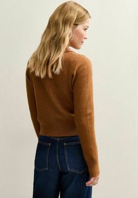 Maglione marrone testurizzato con un design corto e bordo a coste, abbinato a jeans in denim blu scuro. La vista posteriore mostra capelli lunghi fino alle spalle.