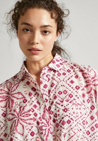 Camisa blanca con patrones geométricos en rosa, cuello puntiagudo y detalle en el bolsillo frontal. Fabricada con un tejido ligero y texturizado.
