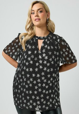 Femme aux longs cheveux blonds portant un chemisier noir avec des motifs d'étoiles argentées et des manches courtes, debout les mains derrière le dos.