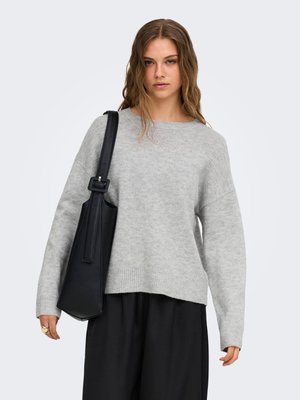 Jeune femme aux cheveux ondulés et lâchés, portant un pull gris et un pantalon noir, portant un grand sac à bandoulière noir.