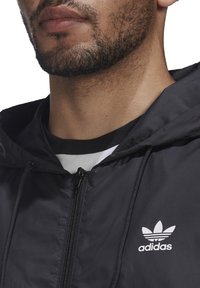 Černá větrovka s předním zipem, kapucí se stahovací šňůrkou a bílým logem Adidas na levé straně hrudi; hladká, lehká látka.
