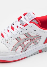 Baskets Asics blanches et rouges avec des bandes de logo grises, des lacets blancs et une doublure intérieure rouge, vues de côté en gros plan.