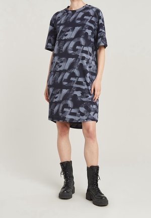 Person iført en løs, mørkegrå knælængde kjole med stor falmet tekst og sorte snørestøvler på en lyse baggrund.