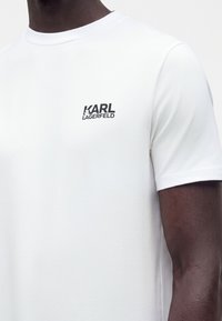 Άνδρας που φοράει απλό λευκό κοντομάνικο T-shirt με μικρό μαύρο λογότυπο "KARL LAGERFELD" στο αριστερό στήθος.