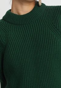 Maglione verde a coste con colletto alto e motivo testurizzato. Presenta leggere linee verticali e polsini stretti.