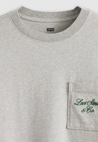 Pelēka kokvilnas kreklu svītra ar priekšējo kabatu. Kabatai ir zaļā izšūta teksta "Levi Strauss & Co.". Maiga auduma tekstūra.