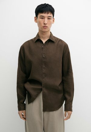 Ung mand står mod en ensfarvet hvid baggrund iført en brun knap-op skjorte og lyse beige bukser.
