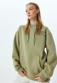 Olijfgroene hoodie met een relaxed fit, textuurstof, trekkoorden, een buidelzak en bedrukte tekst op het linker borstgebied.