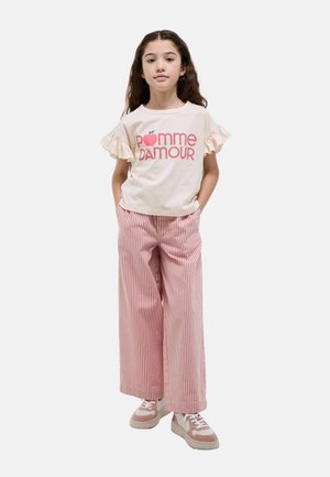 Fille aux longs cheveux portant une chemise crème à manches à volants avec le texte « pomme d'amour », un pantalon large à rayures roses et des baskets roses.