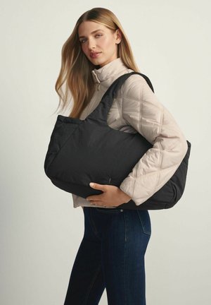 Kobieta o długich blond włosach, ubrana w beżową pikowaną kurtkę i ciemne dżinsy, niesie na ramieniu dużą czarną torbę na pasku.