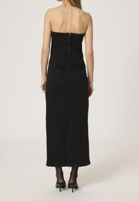 Kvinna som står vänd bort med en axelbandslös svart denim midi-klänning med dragkedja bak, svarta transparenta strumpbyxor och svarta klackskor.