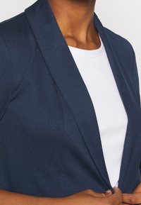 Mörkblå skräddarsydd kavaj med slim fit, med djup notch-lapel och slät tygstruktur över en vit ribbad topp under.