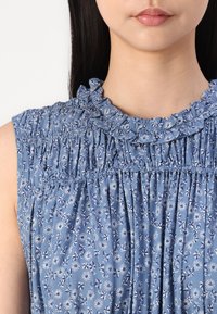 Light blue sleeveless blouse with ruffled neckline, pleats, and floral print in white and navy. Soft fabric with a slightly textured surface.
