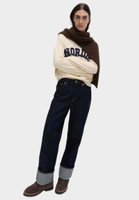 Sudadera crema con la palabra "NORDIC" en texto azul, pantalones vaqueros de mezclilla oscura con puños enrollados y botas de tobillo marrones con una superficie texturizada y costuras.