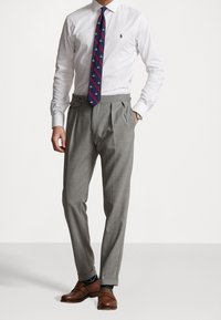 Mies, der ein weißes Hemd, eine dunkelblaue, gemusterte Krawatte, graue, faltige Hosen, braune Loafers und eine Uhr trägt, steht mit einer Hand in der Tasche.