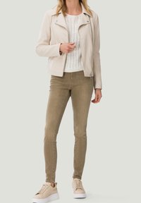 Světlý béžový kožený kabát na zip, nošený přes bílý pletený svetr. Outfit doplňují olivově zelené úzké kalhoty a béžové tenisky na šněrování.