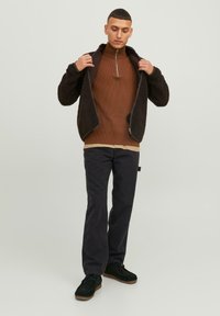 Jersey de cuello alto de rayas marrón debajo de una chaqueta negra de felpa con cremallera, combinado con pantalones oscuros y zapatos negros. Diseño minimalista con elementos funcionales.