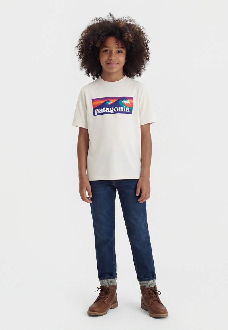 Παιδί με κατσαρά μαλλιά που φοράει λευκό t-shirt με το λογότυπο της Patagonia, μπλε τζιν, γκρι κάλτσες και καφέ μποτάκια με κορδόνια, στέκεται μπροστά σε λευκό φόντο.