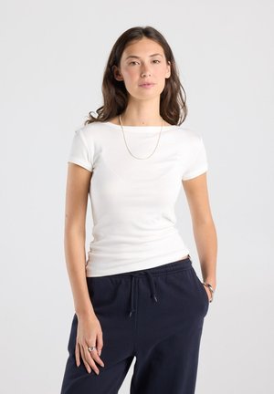 Femme aux cheveux bruns portant une chemise blanche à manches courtes, un pantalon noir et des bijoux minimalistes, debout devant un fond clair uni.