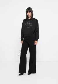 Zwarte sweatshirt met een opvallend grafisch ontwerp op de voorkant, gecombineerd met brede zwarte broek en puntige zwarte schoenen.