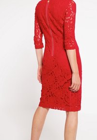 Röd spetsklänning med trekvartsärmar, figurnära form, blommönster och dragkedja i ryggen. Strukturerad yta förhöjer designen.