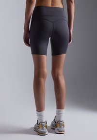 Shorts taille haute gris en tissu lisse et extensible, arrivant à mi-cuisse. Portés avec des chaussettes blanches et des chaussures de sport grises et jaunes.