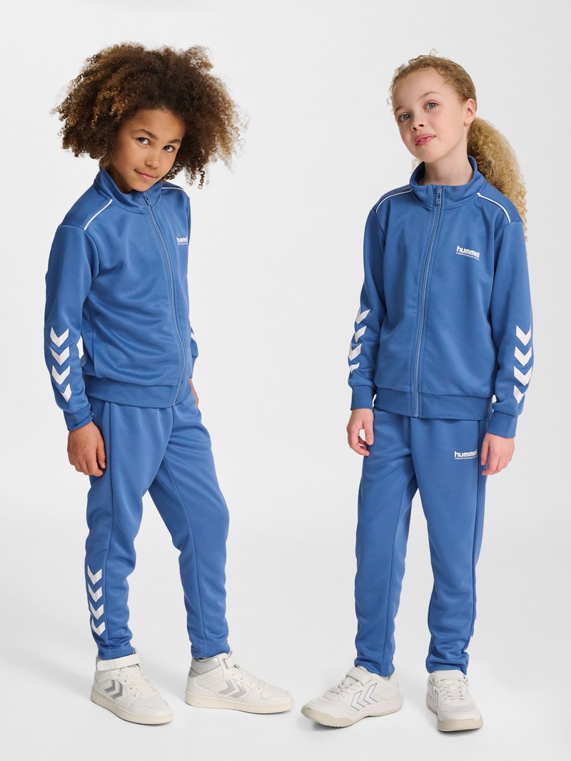 Deux enfants portant des survêtements bleus assortis Hummel et des baskets blanches se tiennent debout devant un fond blanc, face à l’avant dans des poses détendues.