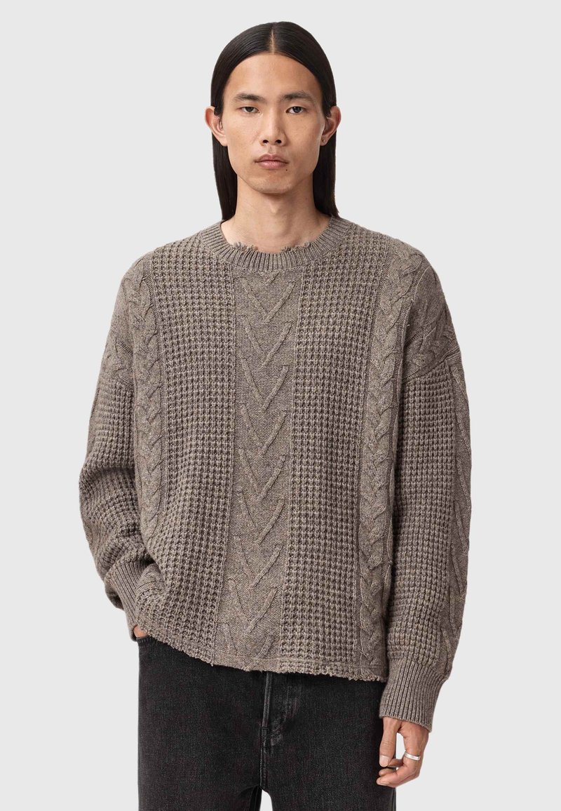 Strikkegenser i lys brun, med kabel- og teksturmønstre, rund hals, og løs passform med frynset kant.