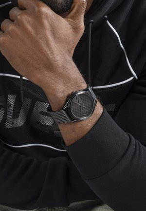 Mann trägt eine schwarze Netz-Armbanduhr mit dem "GUESS"-Logo, stützt sein Kinn auf die Hand und trägt einen schwarzen Kapuzenpullover mit weißen Paspeln.