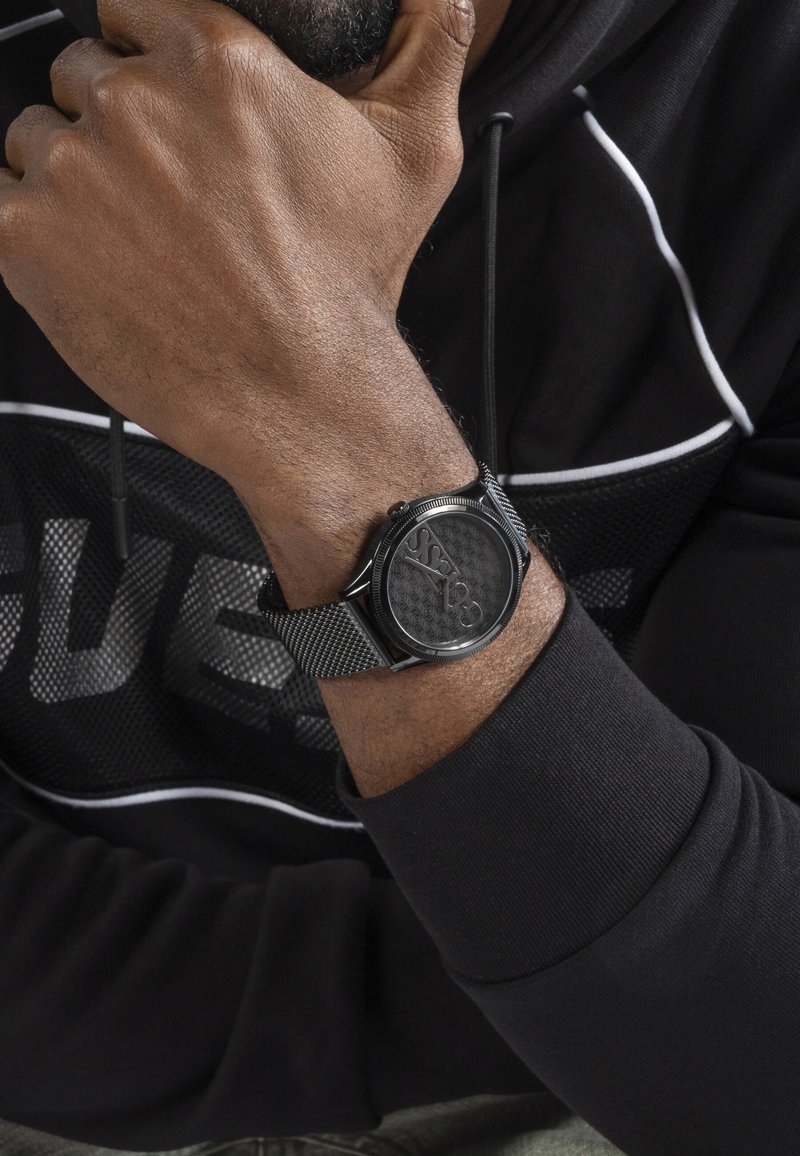 Homme portant une montre noire en maille avec le logo "GUESS", reposant son menton sur sa main, vêtu d'un sweat à capuche noir avec des bordures blanches.