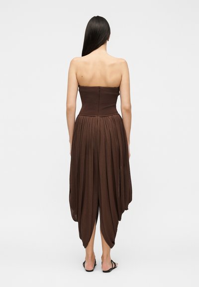 Γυναίκα με μακριά σκούρα μαλλιά φοράει καφέ πλισέ φόρεμα χωρίς τιράντες με ασύμμετρο τελείωμα και μαύρα σανδάλια, στέκεται με την πλάτη προς το λευκό φόντο.