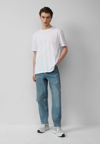 Biała bawełniana koszulka, luźny krój, zestawiona z jasnoniebieskimi, lekko przetartymi dżinsami oraz szaro-białymi sneakersami, stojąca na neutralnym tle.